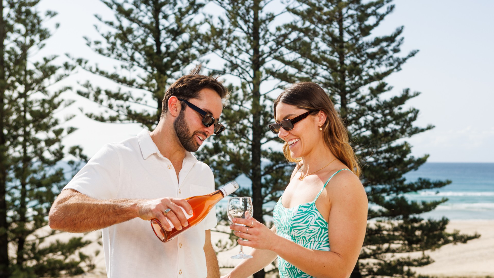 A man is pouring a woman a drink with beach and pine trees in background